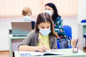 Girl writing at desk wearing facemask with teacher instructing another student at desk behind her, also wearing face masks.