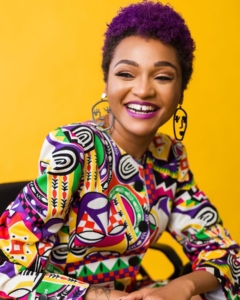 A black woman with a colorful blouse, short purple hair, posing and smiling in front of a gold backdrop.