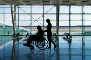A silhouette of a man in a wheelchair being pushed by a woman through an airport terminal.